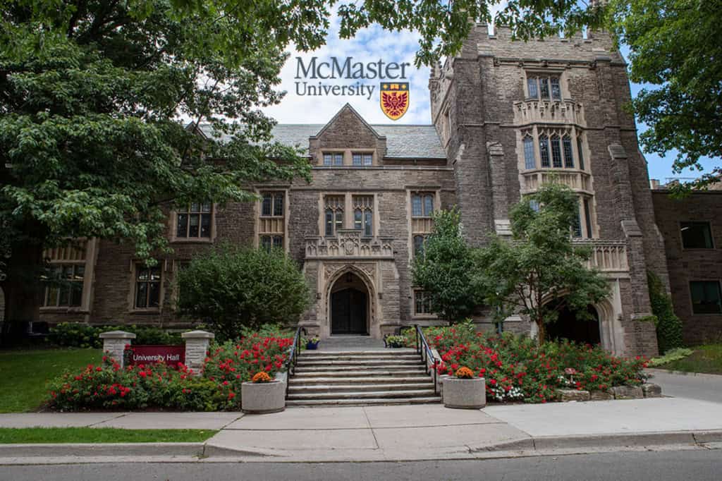 University Hall at McMaster University, a historic stone building with an arched entrance and tall windows surrounded by greenery.