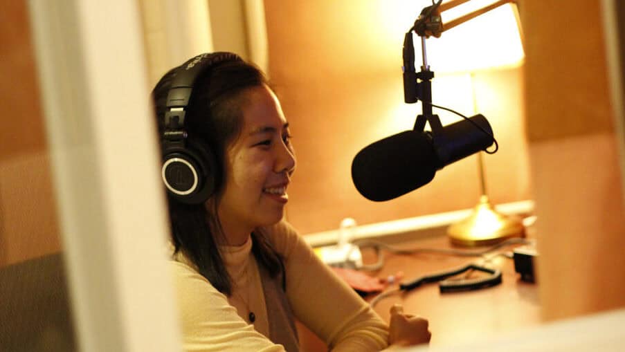A person wearing headphones records at a microphone in the warmly lit audio booth.