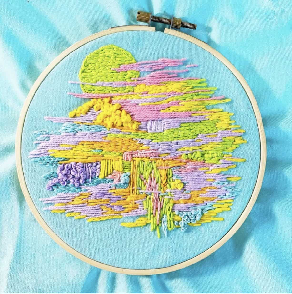 An embroidery hoop displays pastel-coloured thread art, depicting an abstract landscape with a sun and clouds, all stitched on light blue fabric.