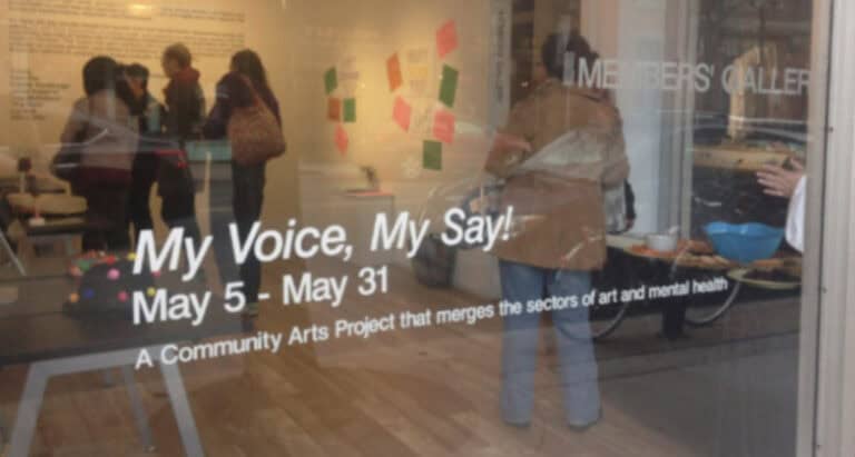 A group of people stand and talk inside Centre[3]'s gallery. Outside on the glass window, white text reads: "My Voice, My Say! May 5 - May 31. A Community Arts Project that merges the sectors of art and mental health".