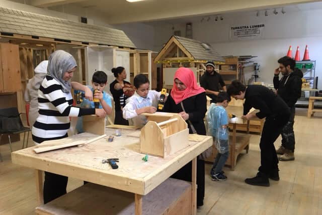 Children and adults work together on woodworking projects in a workshop, using tools like drills and assembling wooden objects at tables. Small wooden houses and construction materials are visible in the background.