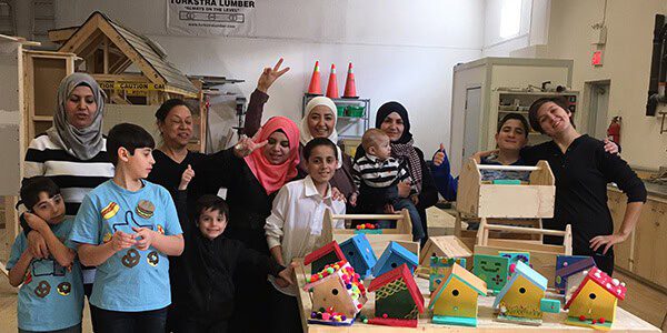 A group of smiling adults and children pose in a workshop, displaying colorful handmade birdhouses and wooden crafts. Some people are holding thumbs up, and a workbench with tools and projects is visible in the background.