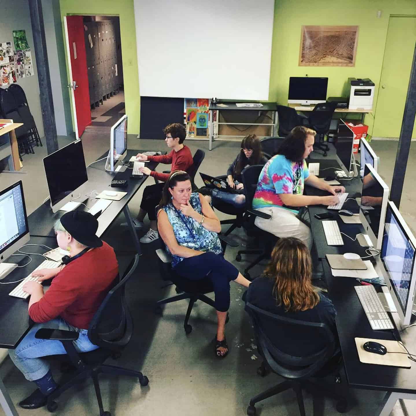 A group of students work at computer workstations Centre[3]'s computer lab with green walls. Some students are focused on their screens, while the teacher in the centre sits with a thoughtful expression.
