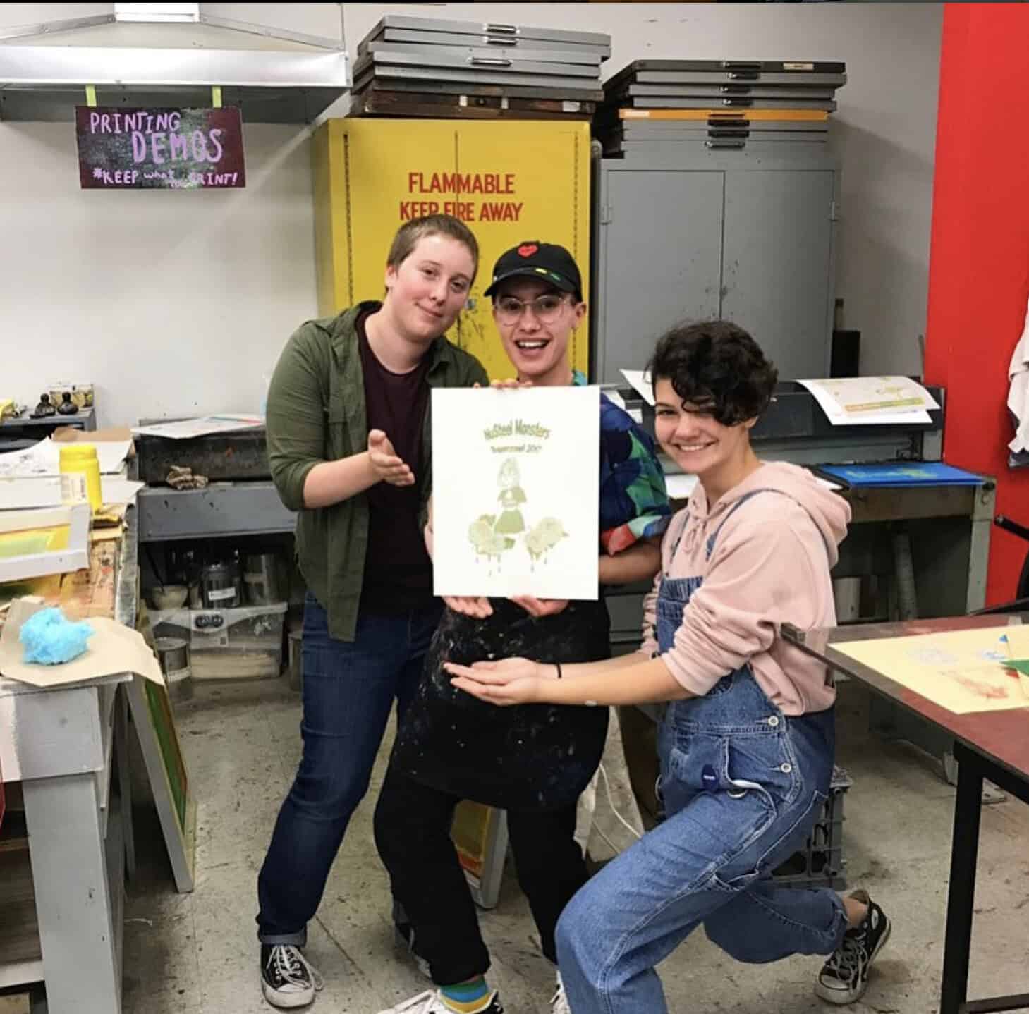 Three young people smiling and posing in Centre[3]'s Traditional Printmaking Studio, holding up a print with a green design. Art materials, tables, and a yellow cabinet are visible in the background.