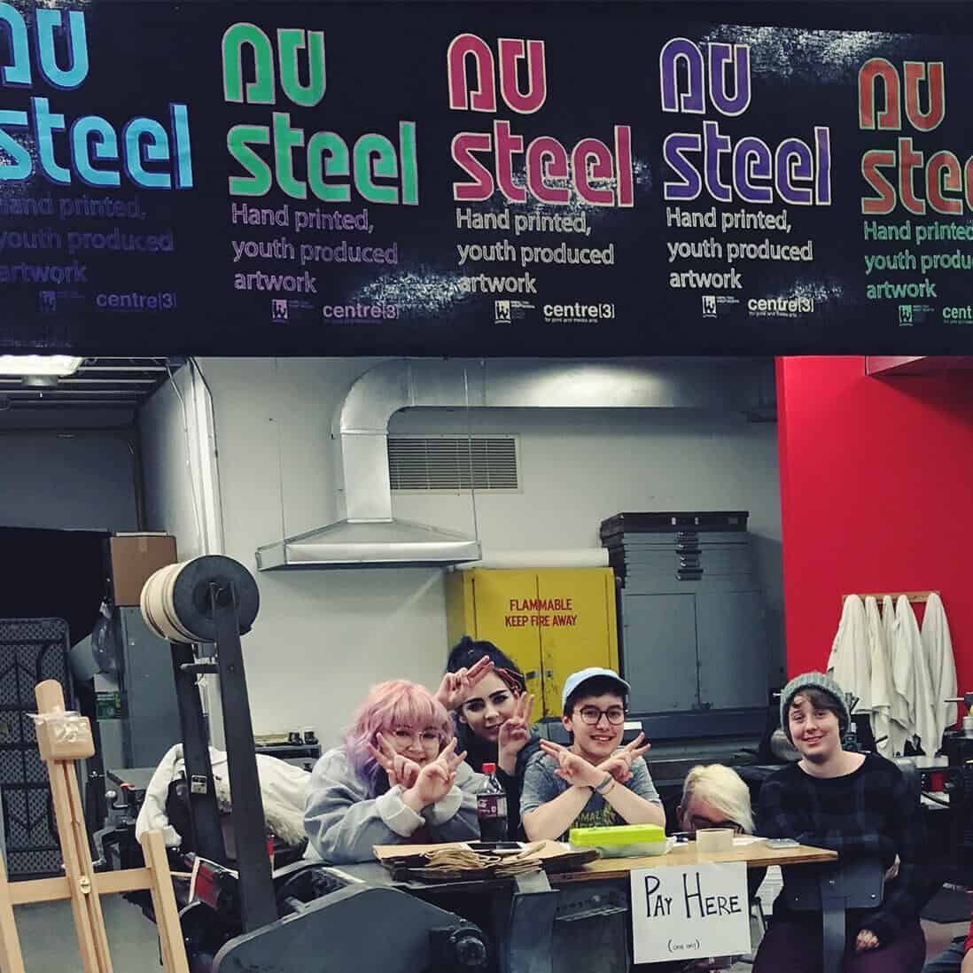 Four smiling young people pose behind a table with a “Pay Here” sign in an art studio. A bright “NuSteel: Hand printed, youth produced artwork” sign hangs above them. Art supplies fill the background.
