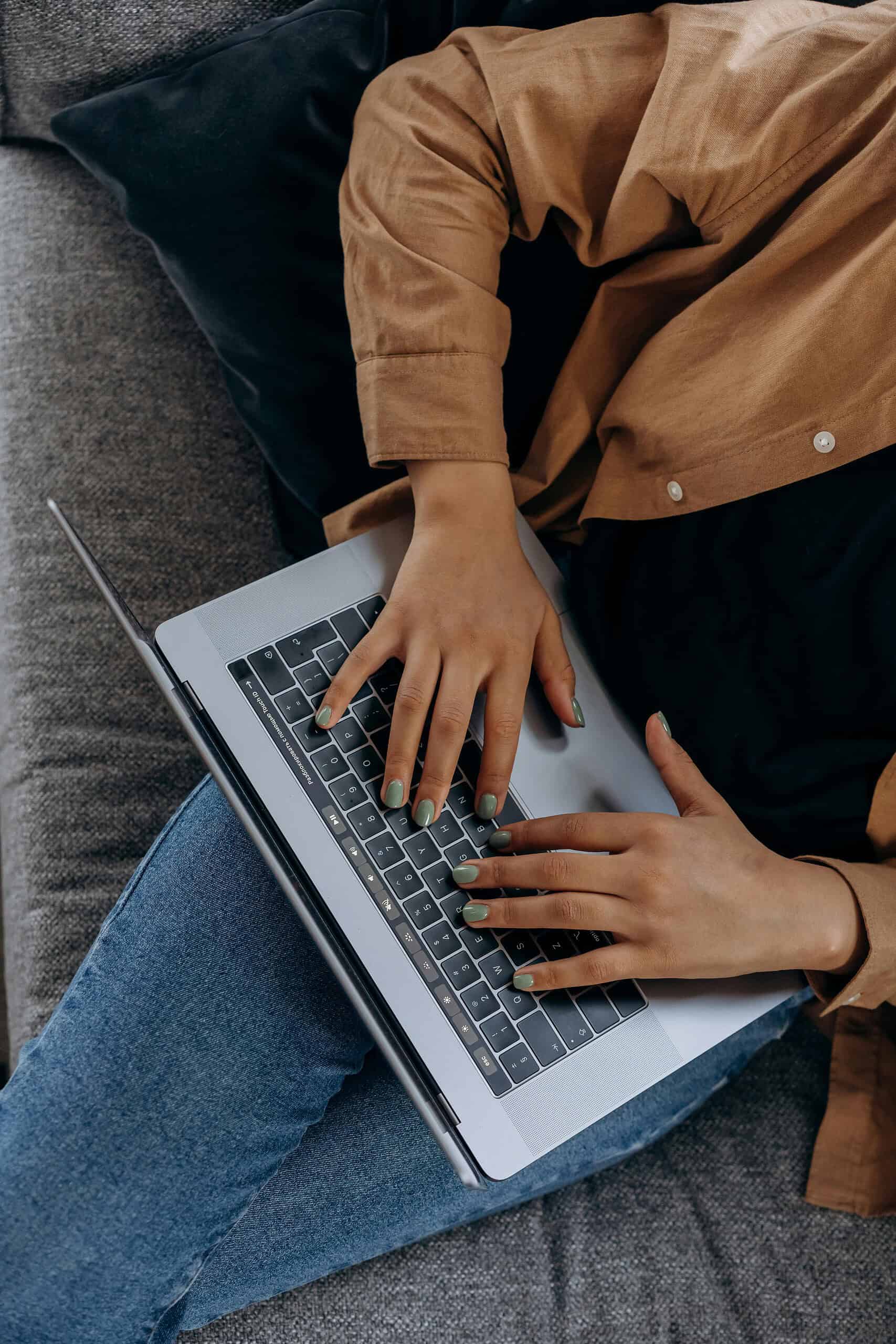 A person in a brown jacket and jeans sits on a grey couch, typing on a laptop computer. The image, shot from above, highlights their hands as they navigate the keyboard.