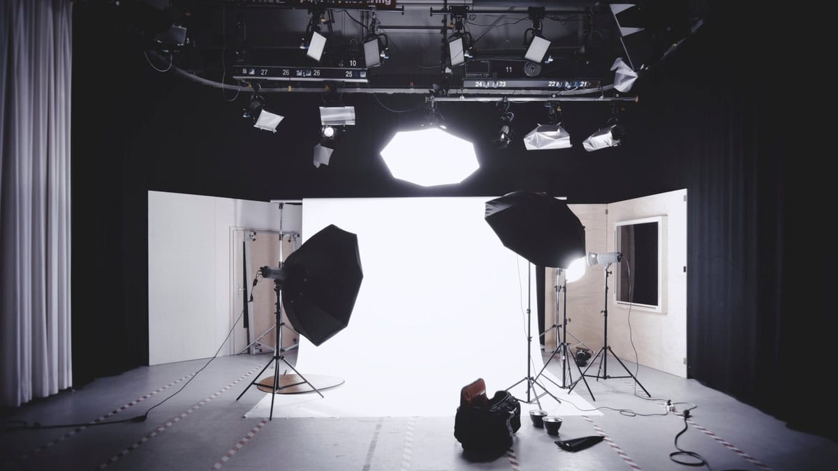 A professional photography studio setup with large lights, umbrellas, tripods, and a bright white backdrop, surrounded by black curtains and light stands.