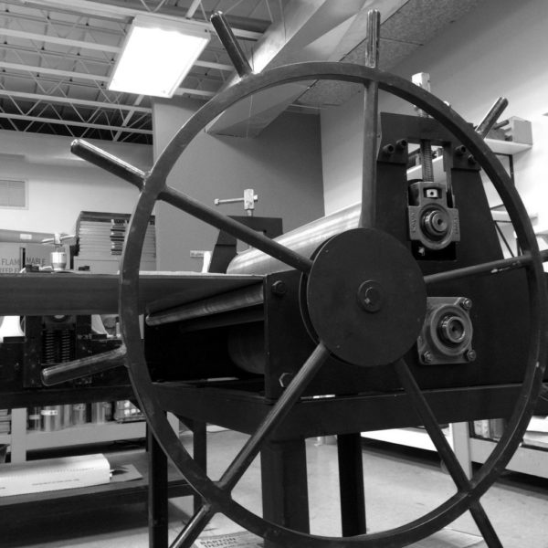 A large wheel and roller of a printing press. The black and white image highlights the metal structure and art studio setting, with shelves and fluorescent lights visible in the background.