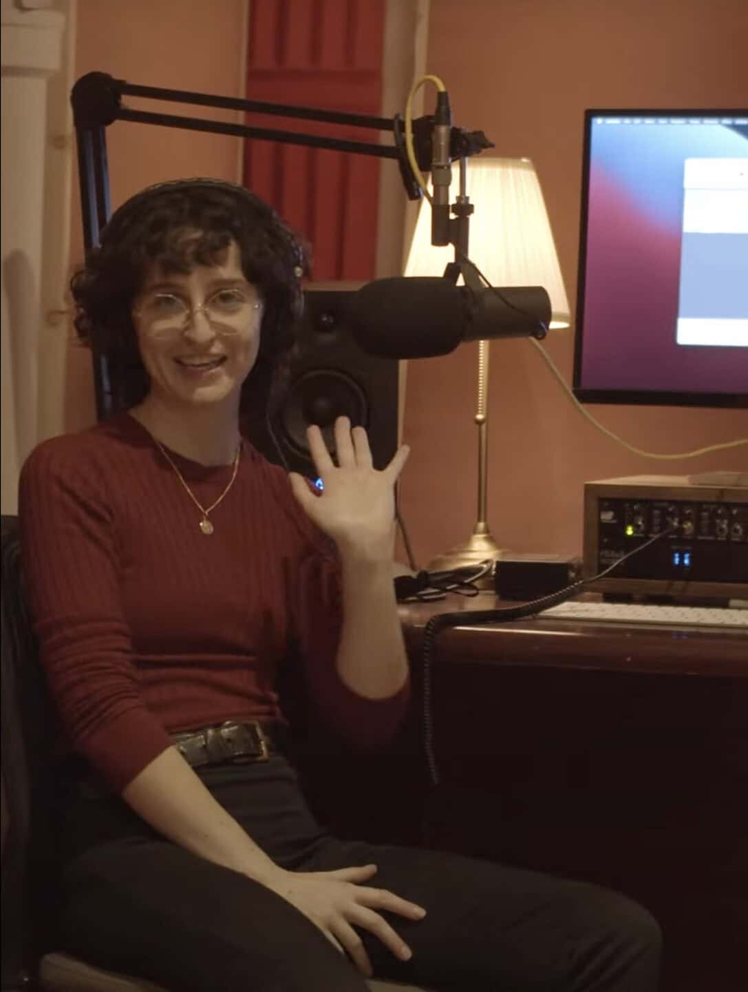 Rebecca Casalino, with short curly hair, glasses, and over-ear headphones, sits at a desk with a microphone, computer, and lamp, smiling and waving at the camera in the Media Arts Studio.