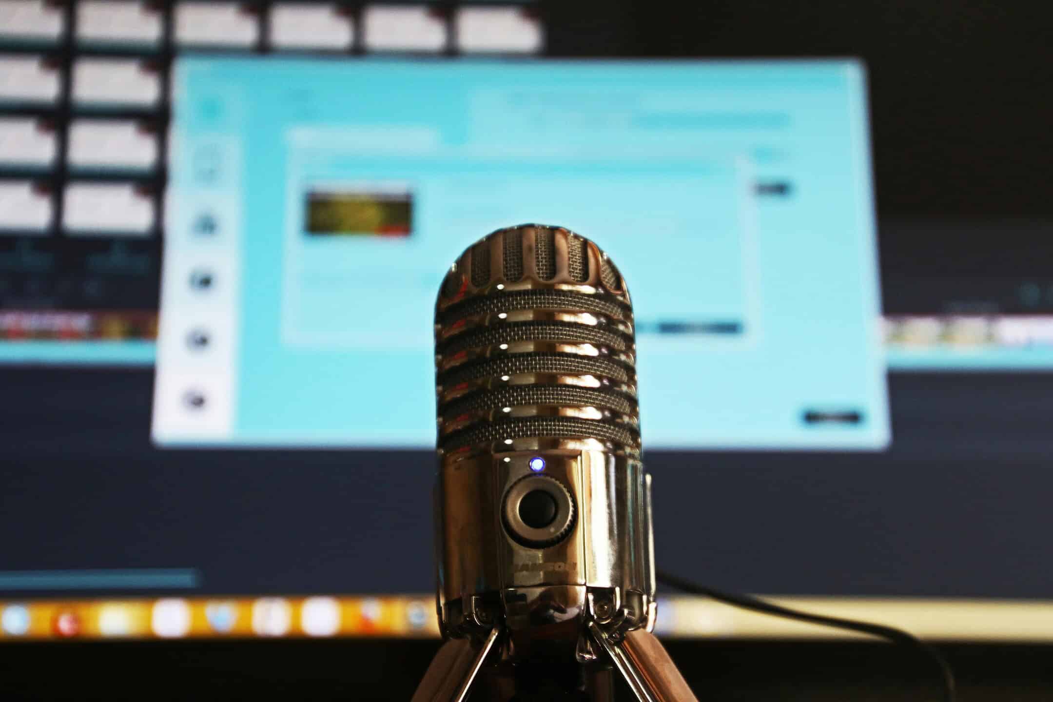 A close-up of a silver microphone on a stand, with a blurred computer screen displaying software and video editing timelines in the background.