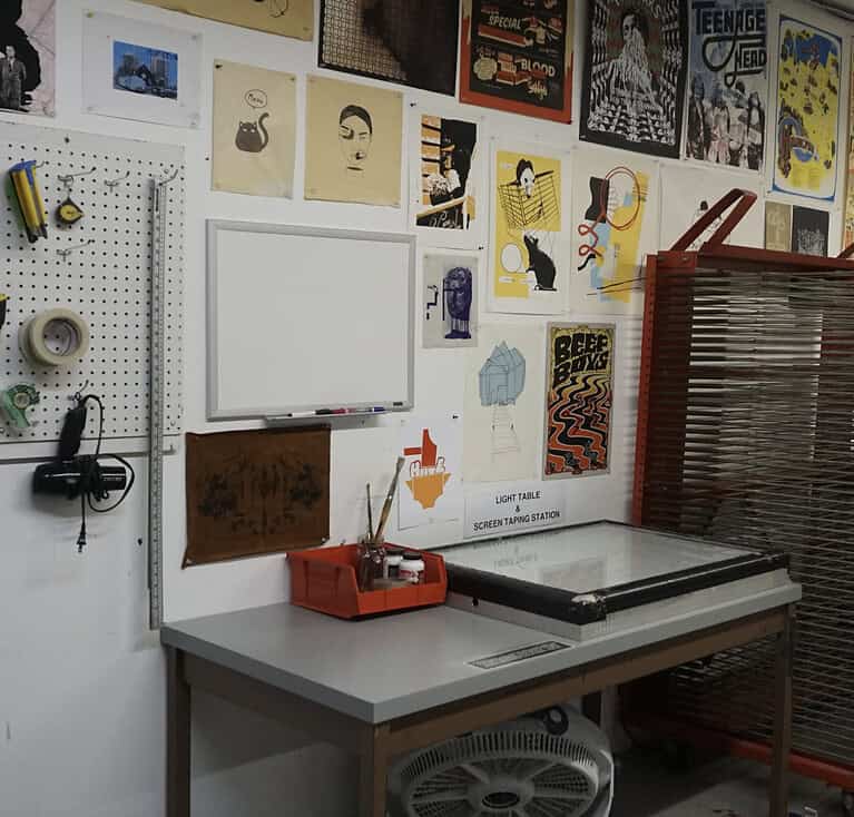 A work station by a studio wall with various colourful posters, a whiteboard, and a pegboard holding tools above a worktable. A light table and fan sit beneath the table beside a drying rack.