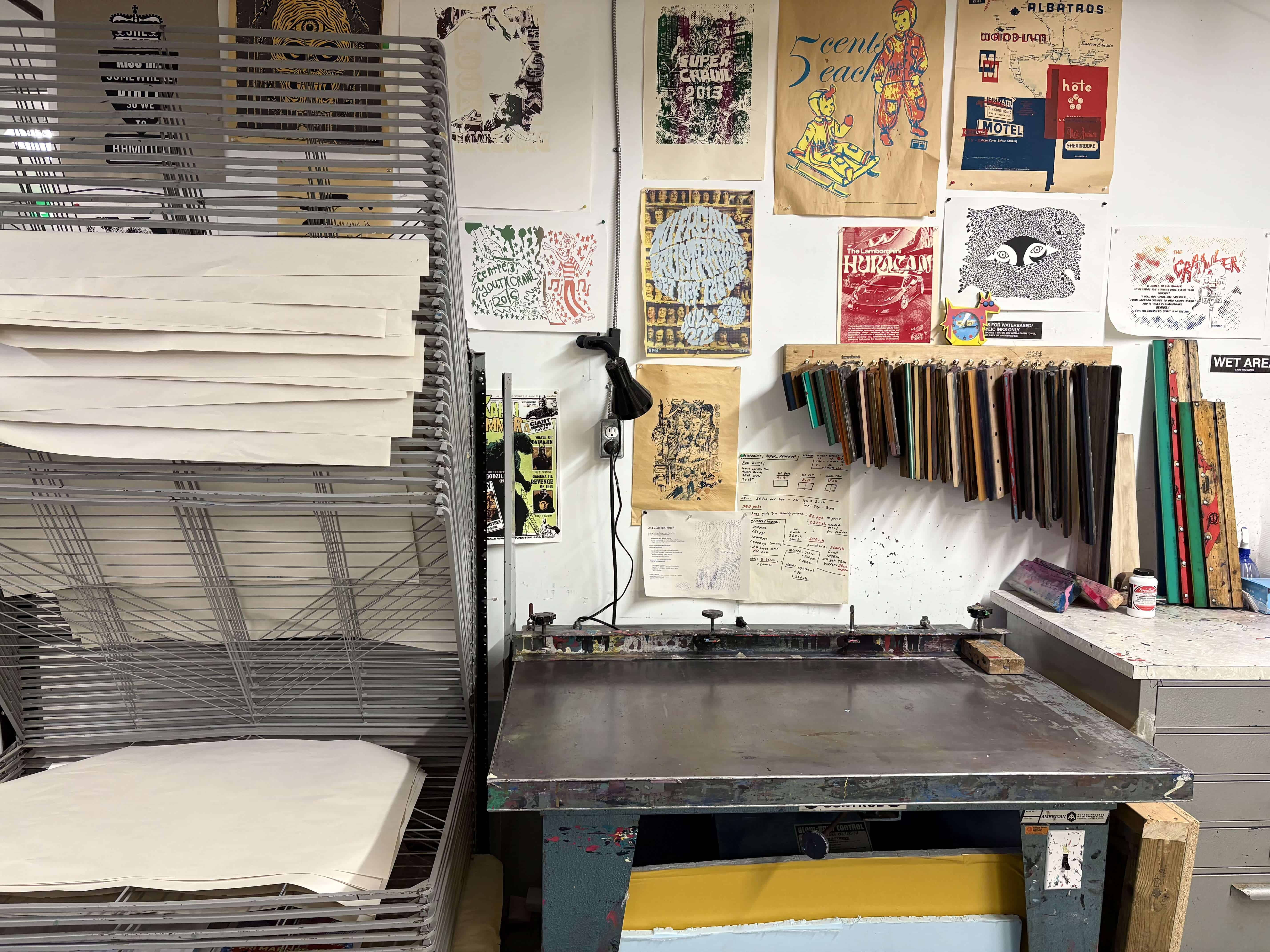 Centre[3]'s Screen Printing Studio with a metal drying rack stacked with paper, a large flat worktable, squeegees hanging on the wall, and colourful prints and posters displayed above the workspace.