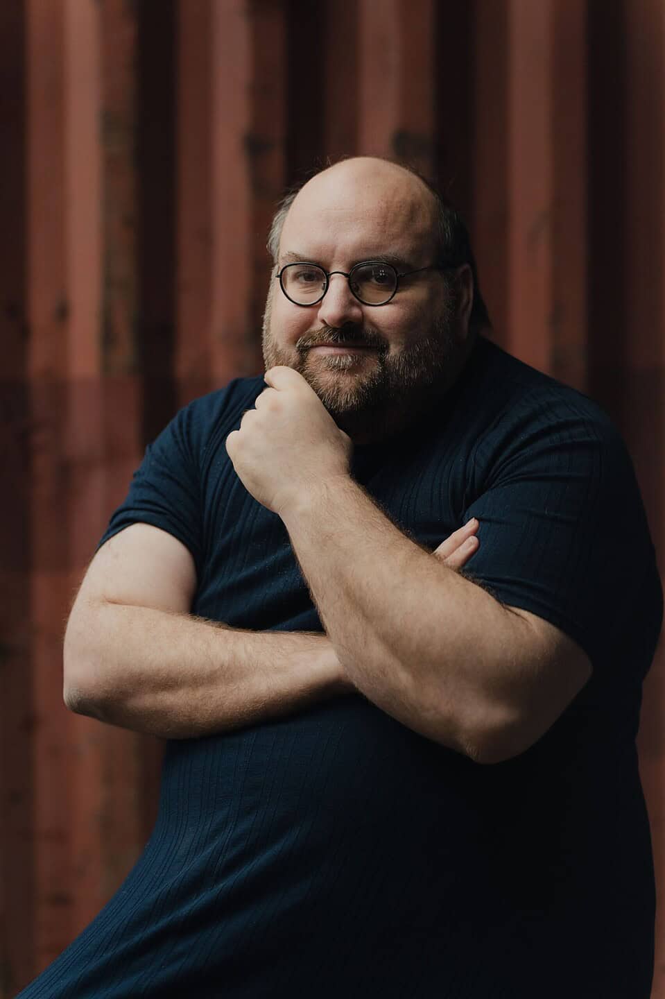 Portrait of Steacy Easton with glasses and a beard posing with arms crossed against a warm-toned textured background