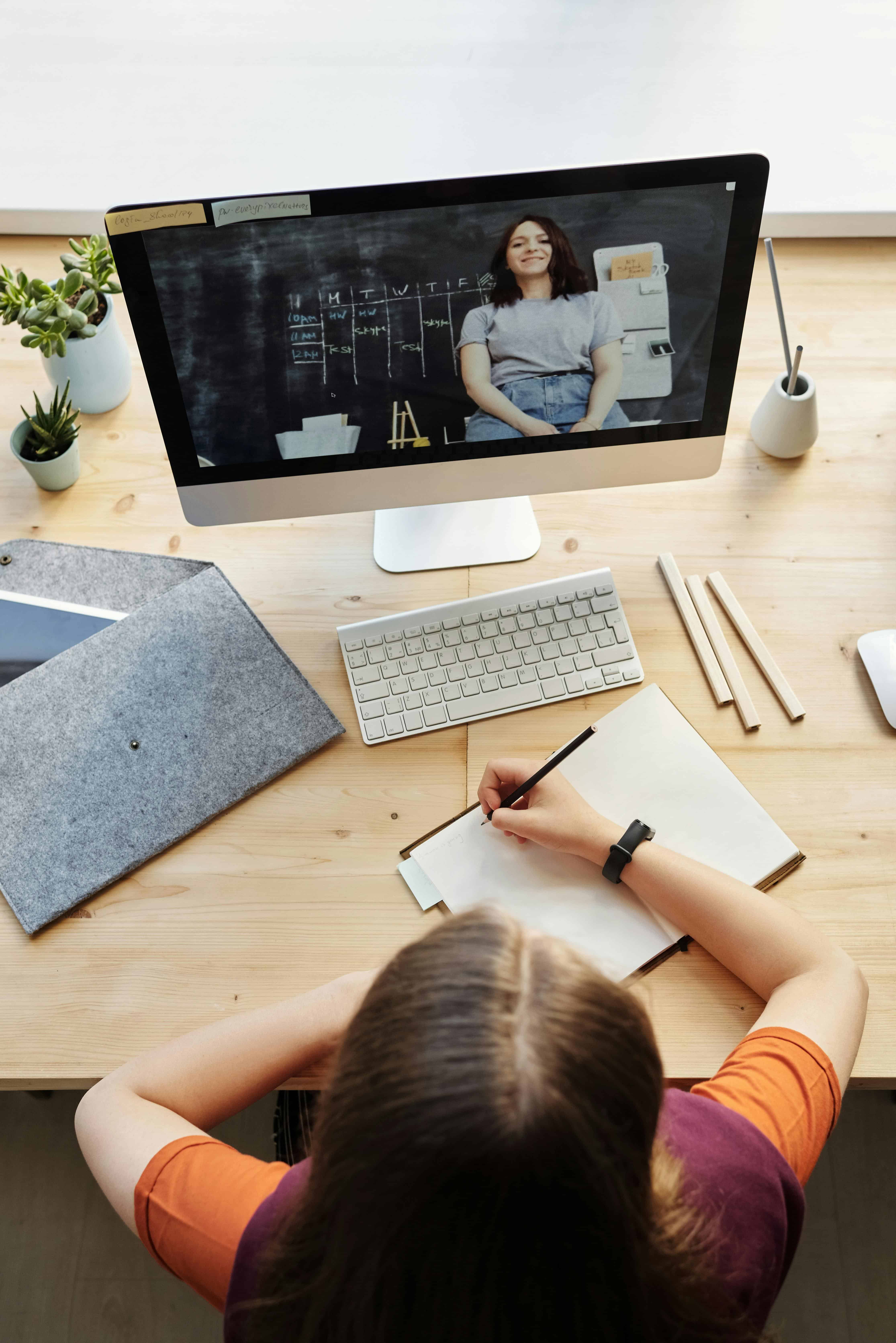 A youth sits at a desk taking notes on a notepad while attending a video call with a person on the computer screen. The desk holds a keyboard, pens, plants, and folders.