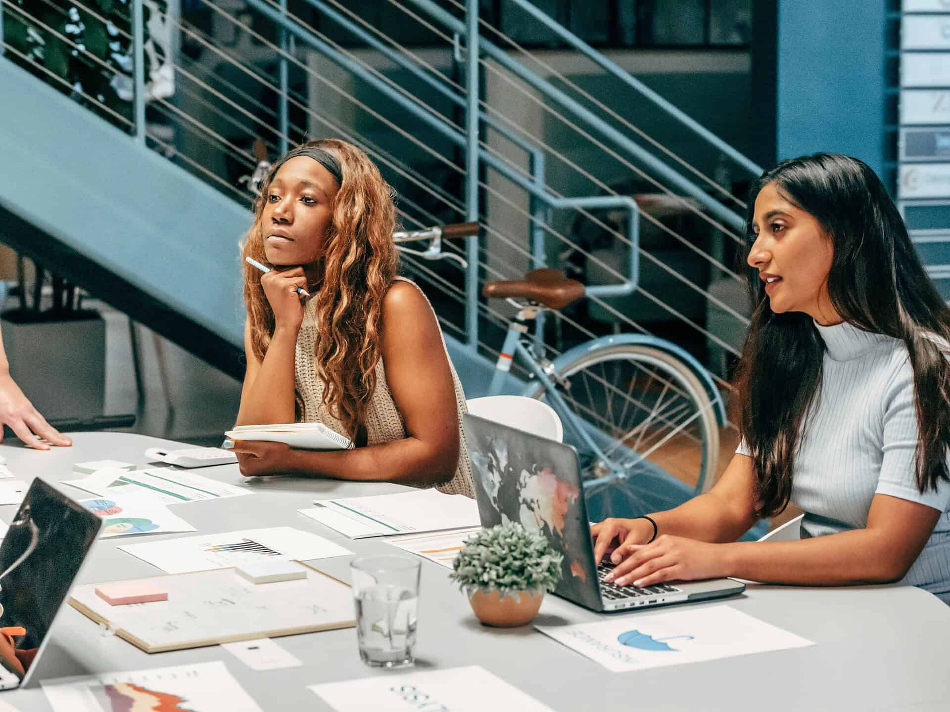 Two young people sit at a table with documents and a laptop, appearing focused during a meeting. Behind them are a staircase, a bicycle, and a blue wall.