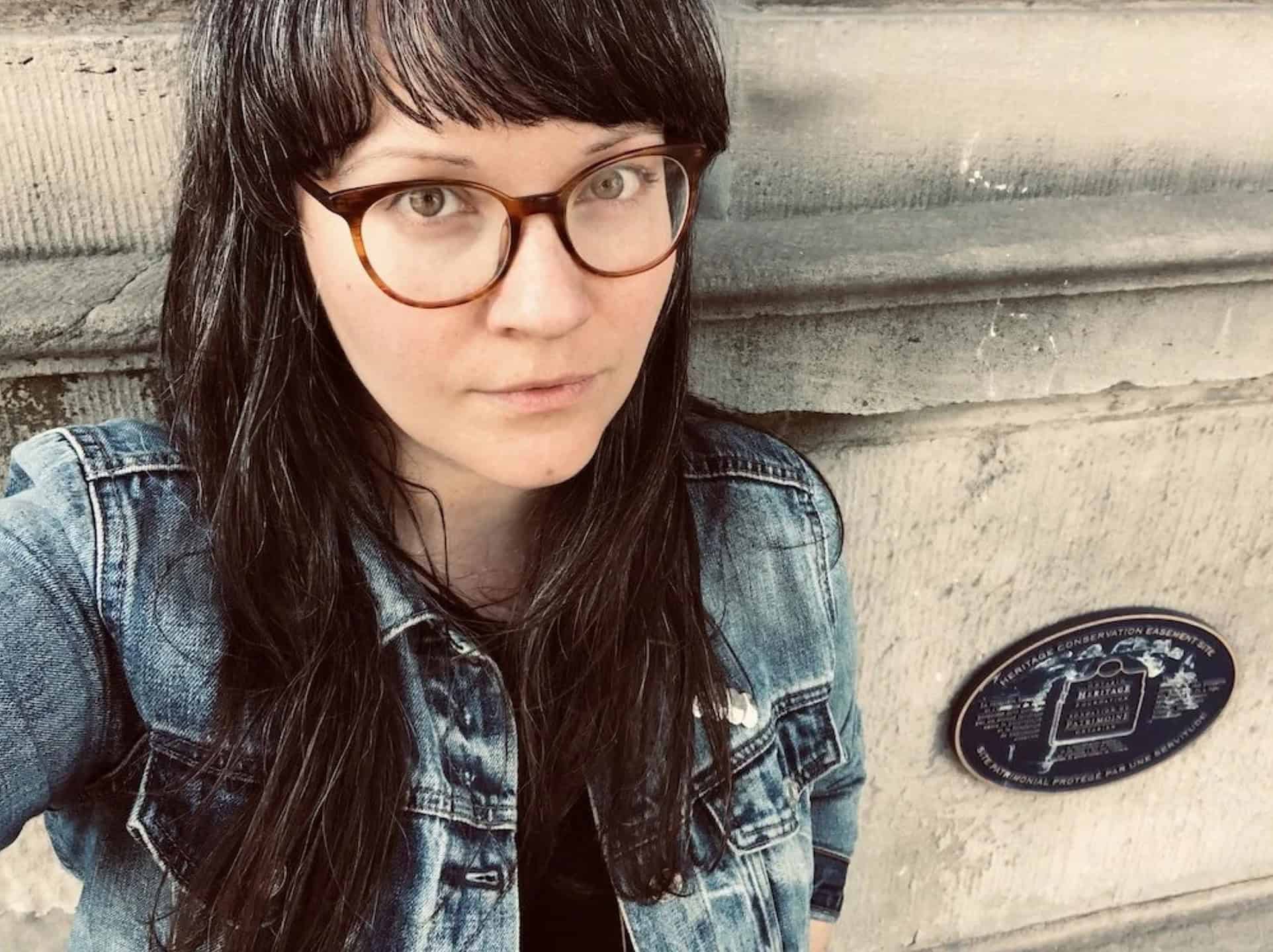 Portrait of Tara Bursey with dark hair and glasses wearing a denim jacket, standing beside a stone wall with a heritage plaque