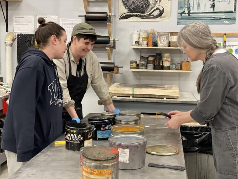 Three people stand around a worktable in the Traditional Printmaking Studio with large cans of ink on the table. One person stirs a container of ink while the others observe and smile. Art supplies and prints line the shelves and walls in the background.