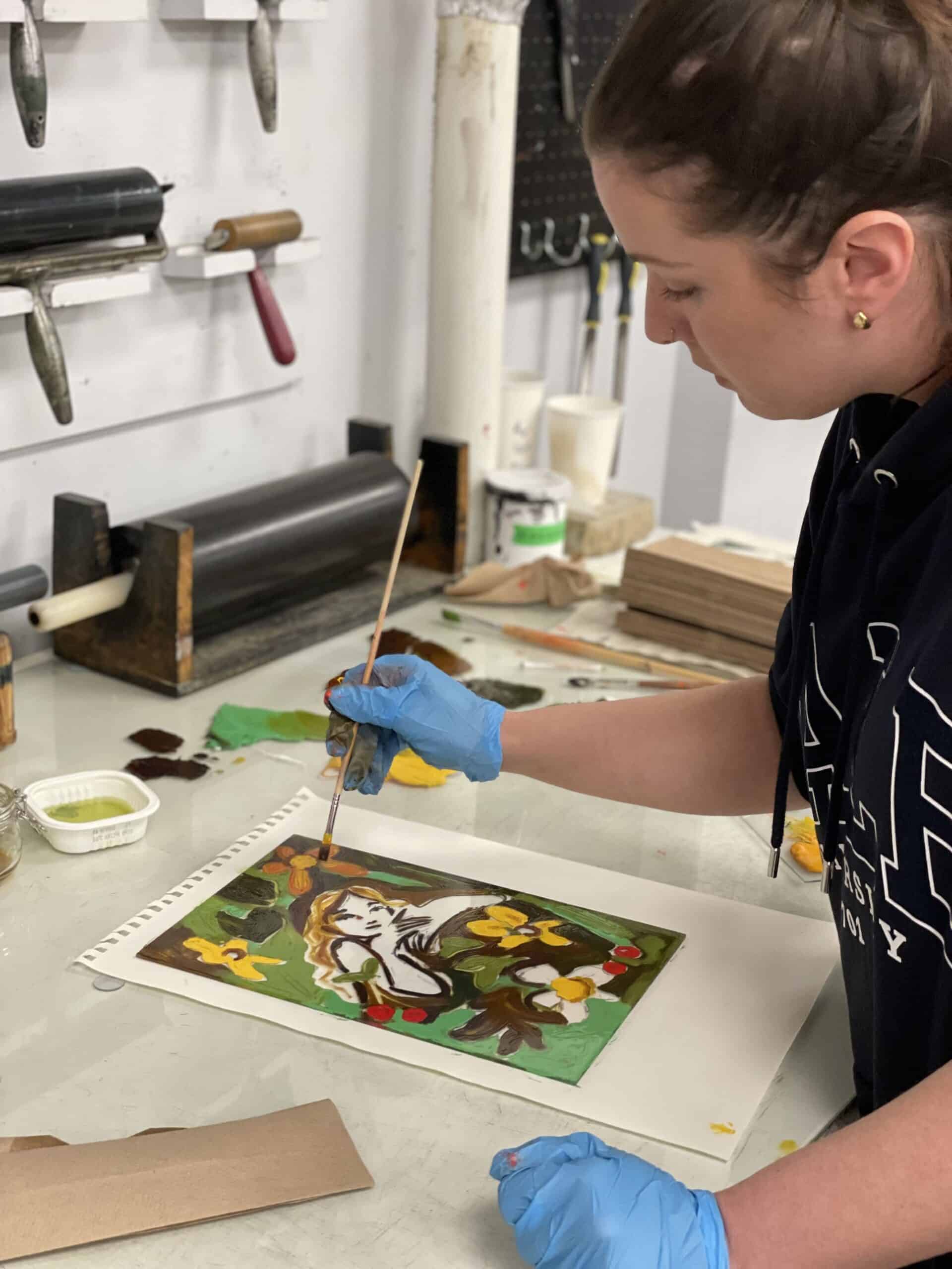 An artist wearing blue gloves uses a brush to apply colour to an artwork depicting a stylized face with flowers. They work at a table with art supplies surrounding them in Centre[3]'s Traditional Printmaking Studio.