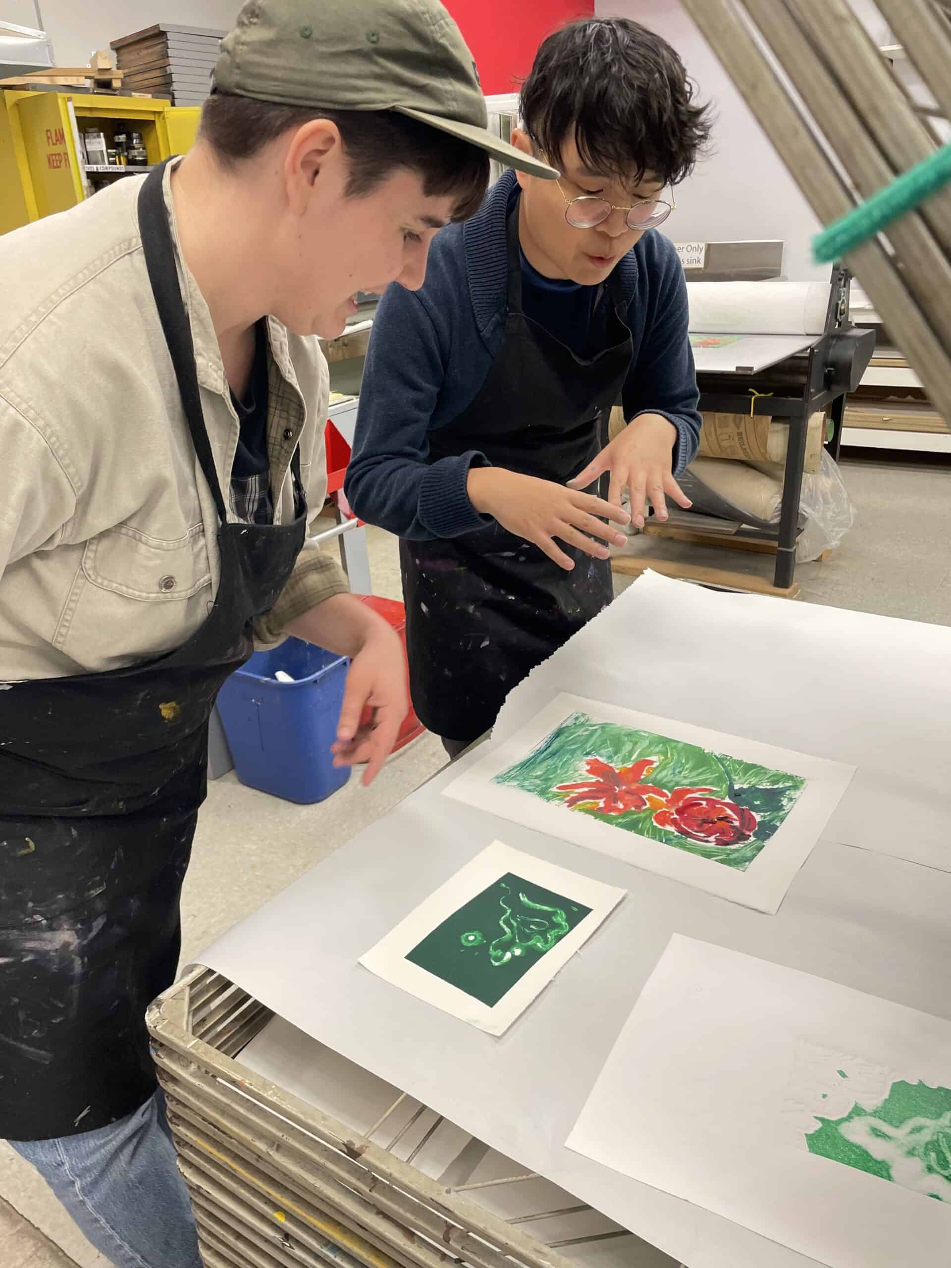 Two people in paint-spattered aprons look at colourful prints drying on a rack in Centre[3]'s Traditional Printmaking Studio. One person gestures while talking, and the other smiles as they observe the artwork. Art supplies and shelves are visible in the background.