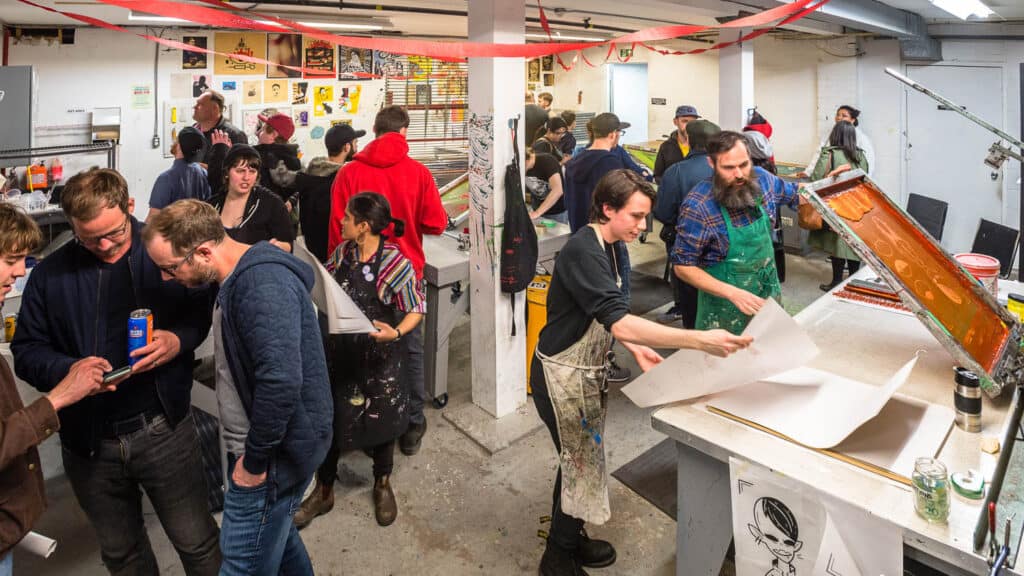 A busy Screen Printing Studio at Centre[3] filled with people socializing and working; some are gathered in groups talking, while others are focused on screen printing and preparing art materials at large tables.