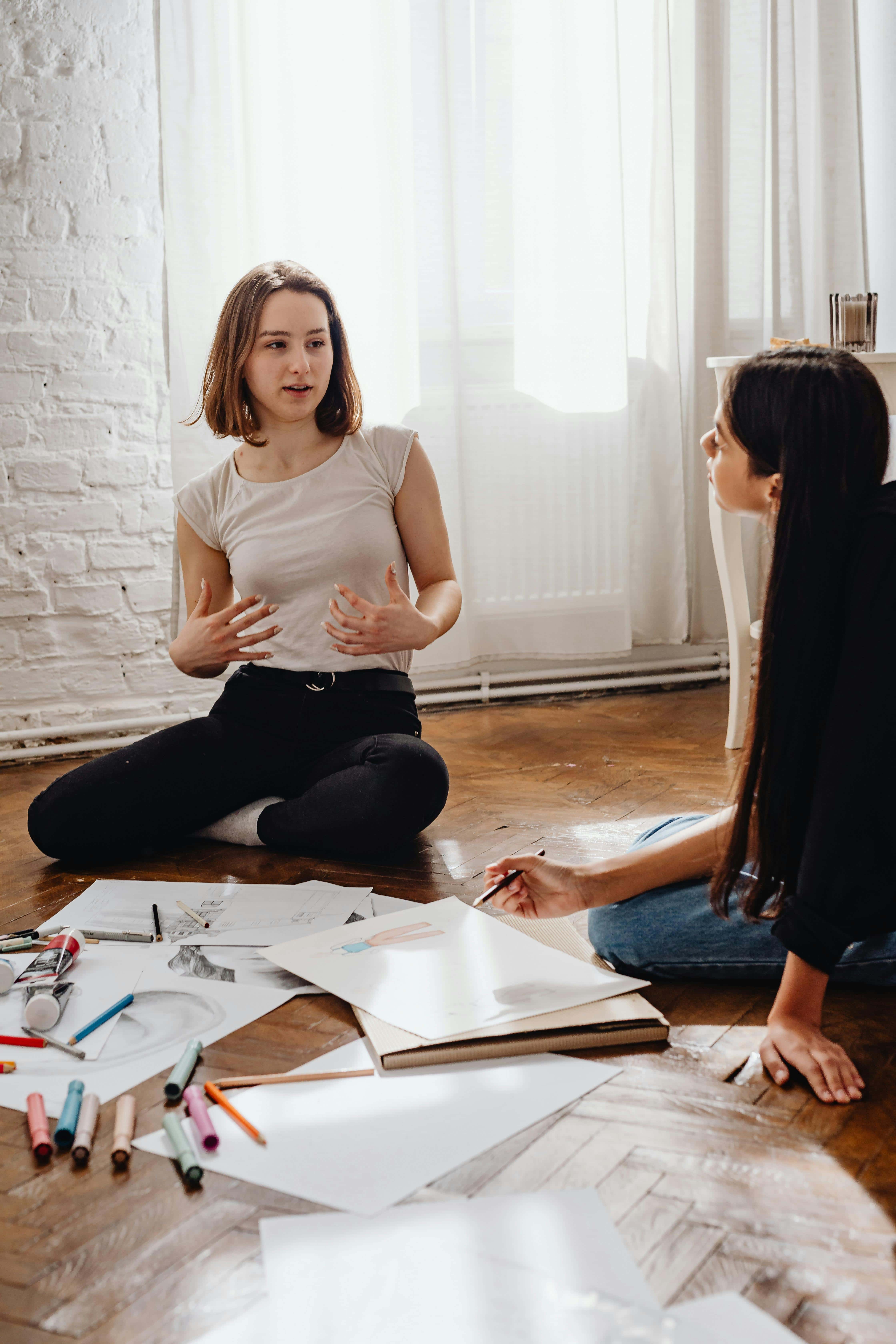 Two young people sit on a wooden floor, surrounded by markers and sketches, having a conversation. One gestures with her hands while the other listens, poised with a pencil. Sunlight filters through white curtains behind them.