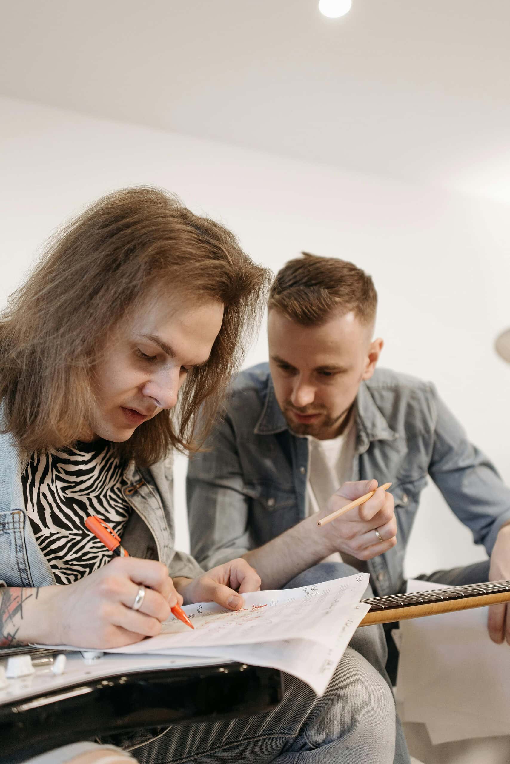 Two people sit together, one is focused on writing notes with a red pen while the other watches and holds a pencil, both appearing engaged and thoughtful.