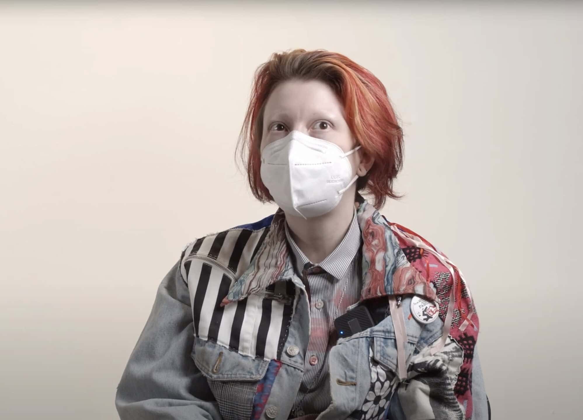 Venus Underhill with short red hair wearing a face mask and a patchwork denim jacket sits against a plain background, looking upward