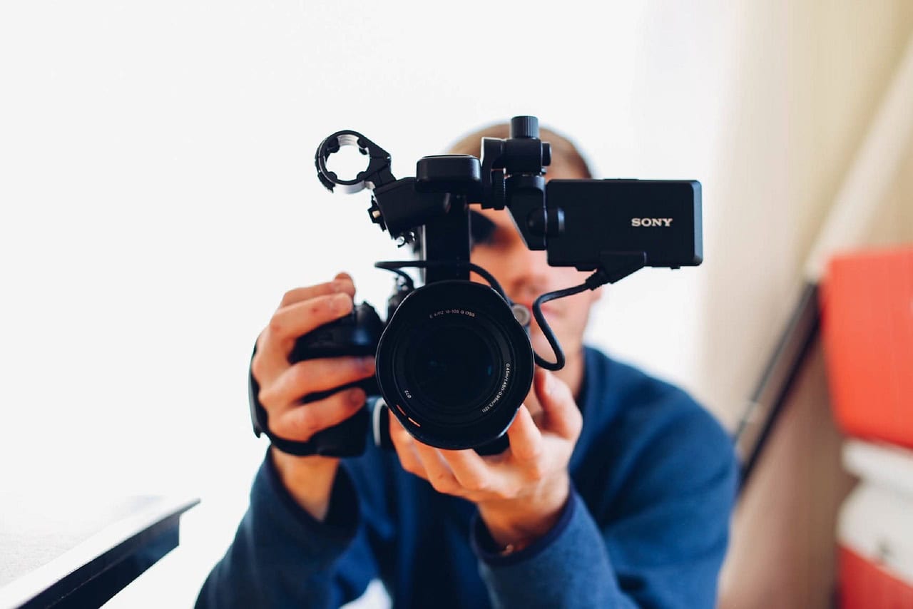 A person wearing a blue top holds a professional Sony video camera, pointing it directly at the viewer. The softly blurred background with light tones draws focus to the camera.