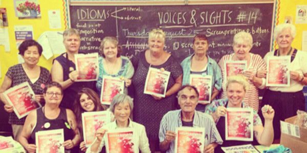 A group in the Voices and Sights seniors program smiling and holding up colourful printed papers in a bright classroom.