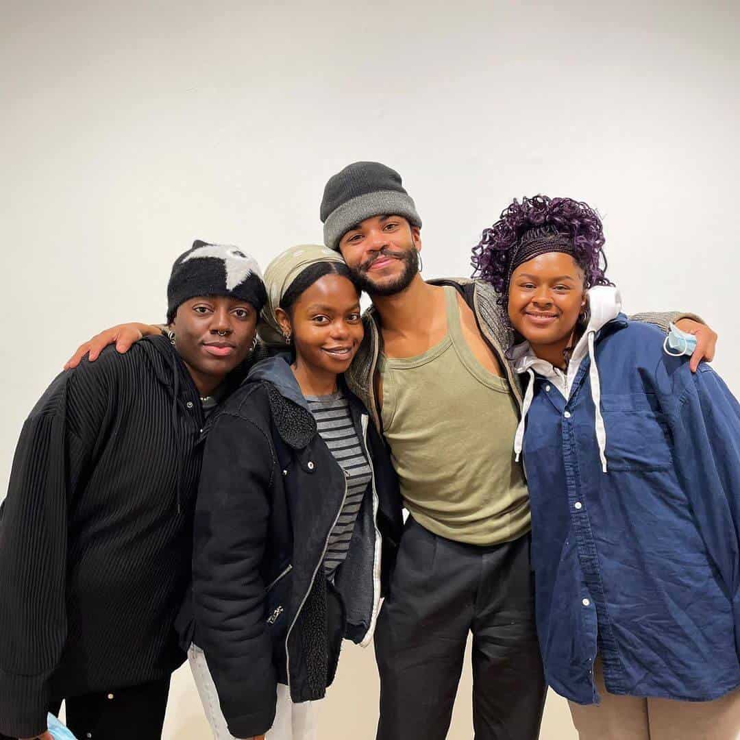 Four people stand close together smiling at the camera with arms around each other against a plain wall