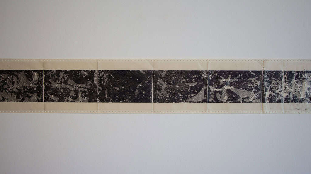 Rectangular paper strip from the exhibition "Work on Paper" with perforated edges, featuring a black and white abstract, marbled pattern printed in the centre. The paper is attached to a white wall.