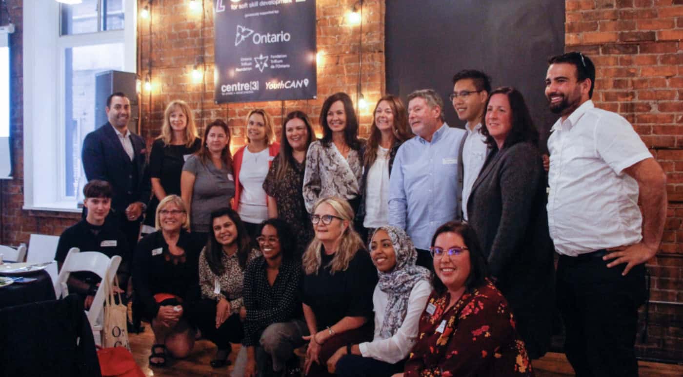 A group of twenty people, diverse in age, gender, and ethnicity, pose together indoors in front of a brick wall. Most are smiling, and some are seated while others stand behind them.