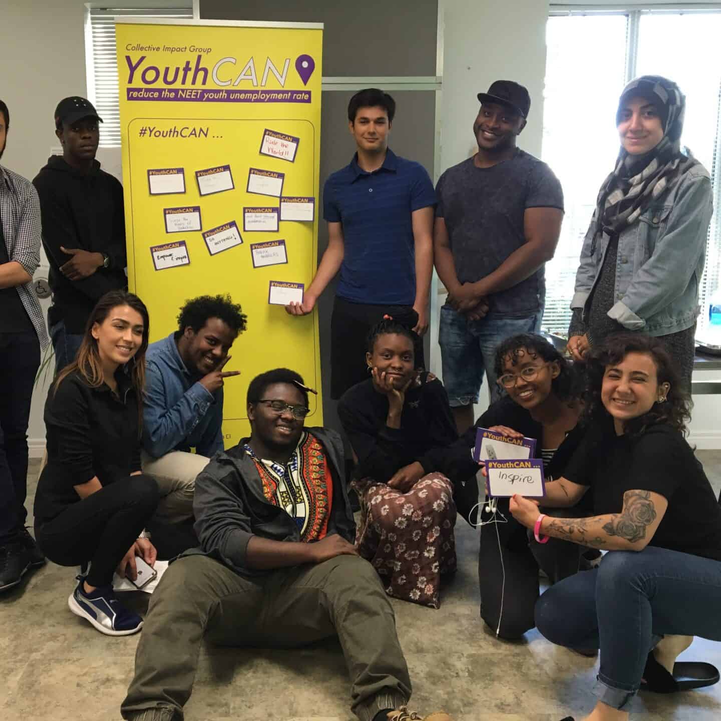 A diverse group of thirteen young adults pose and smile together indoors in front of a yellow YouthCAN banner about reducing youth unemployment. Some are standing, others kneeling or sitting on the floor.