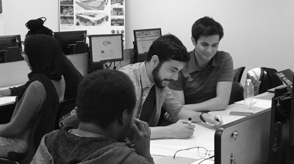 Four students work together at desks with computers in a classroom; two are collaborating closely on a project, while two others are focused on their own work. The scene captures a collaborative learning environment.