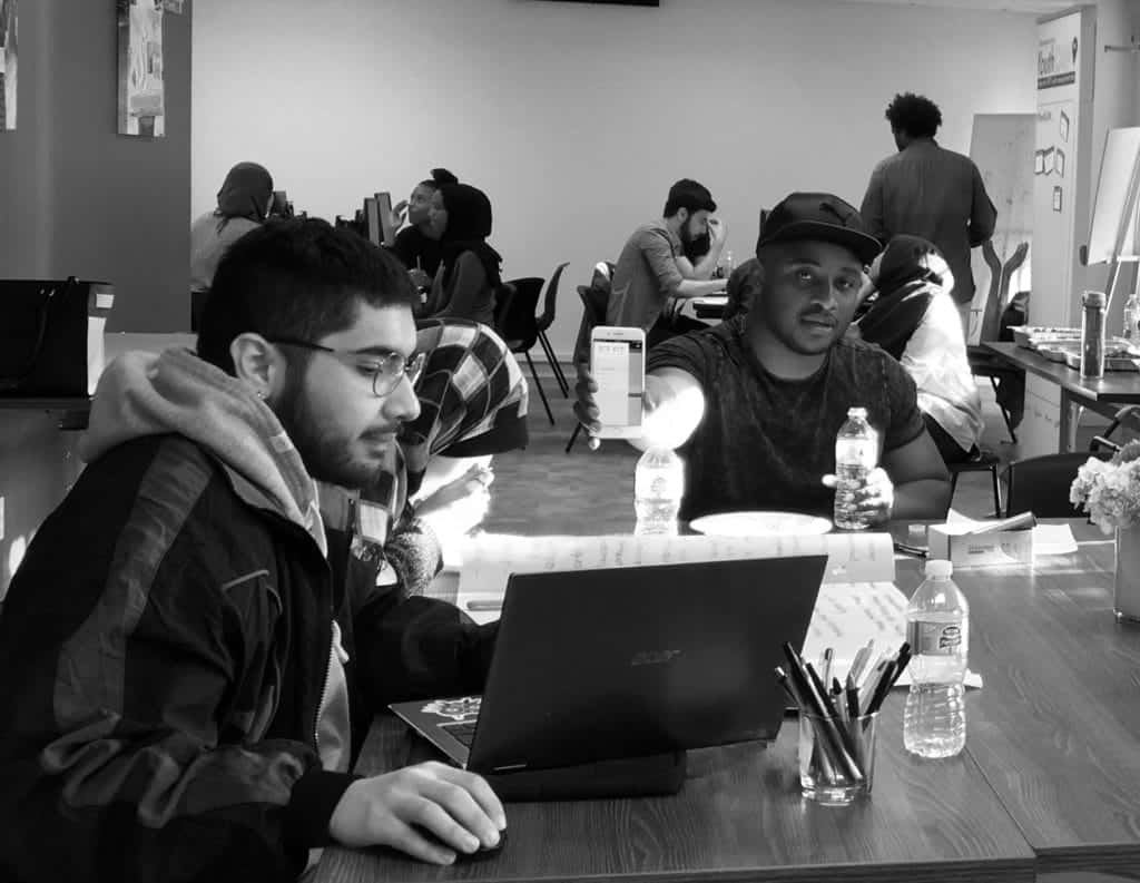Black and white photo of several people working and talking at tables in a shared indoor space. One man holds up a bottle, while others focus on laptops and papers. Water bottles, pencils, and flowers are on the table.