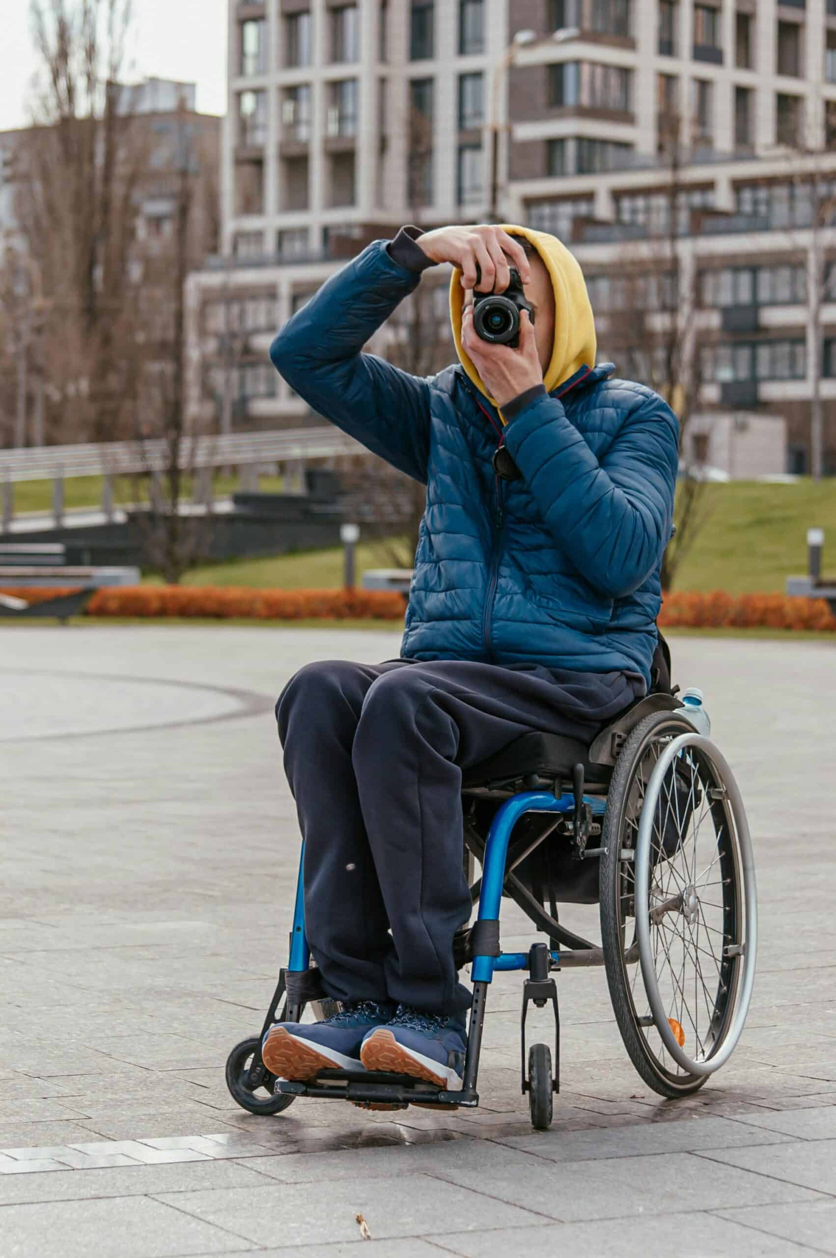 A person in a wheelchair wearing a blue jacket and yellow hoodie takes a photo with a camera outdoors near the city centre.