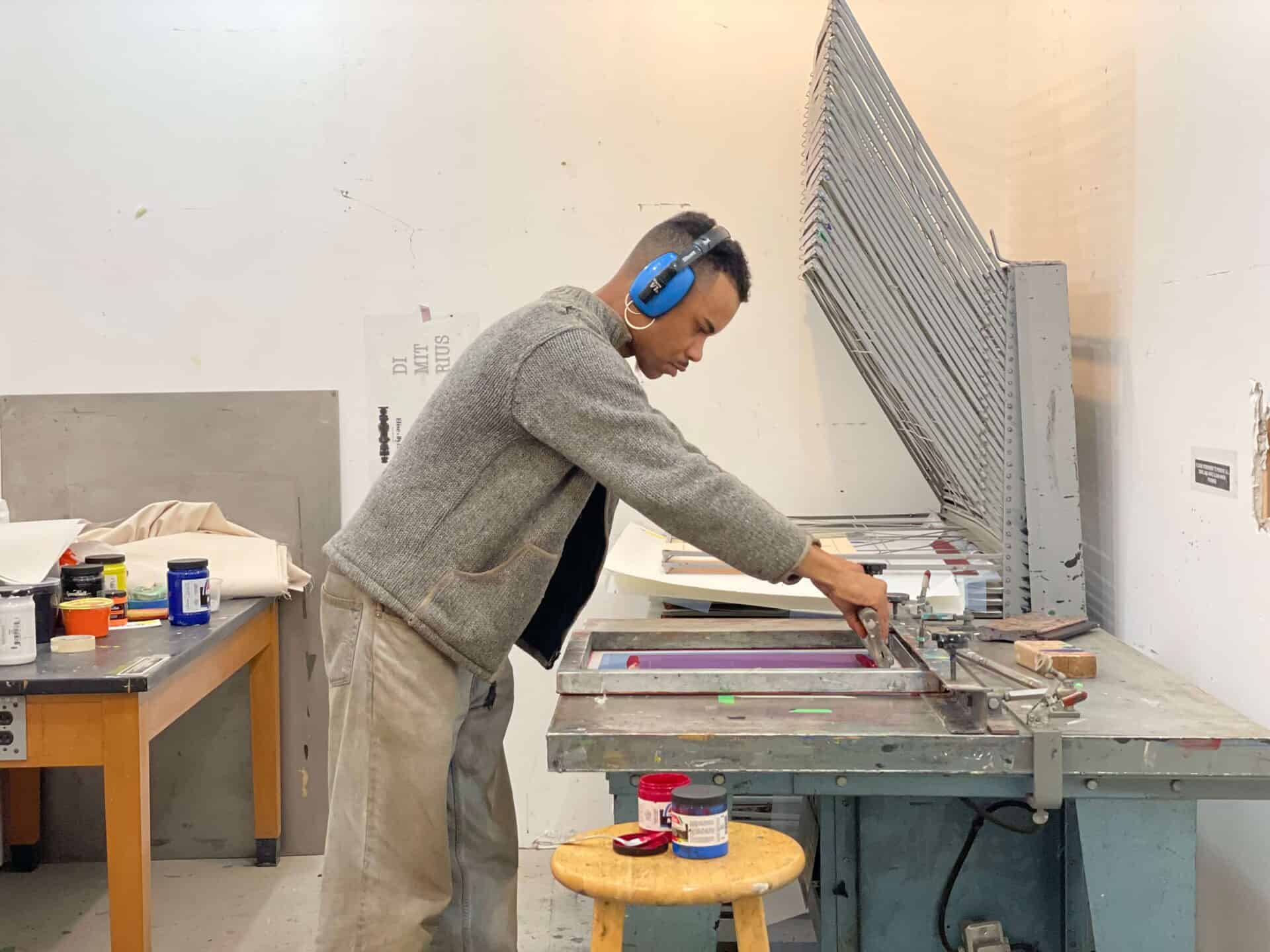 An artist wearing blue headphones and a grey jacket operates screen printing equipment in the studio, with ink containers and supplies on a nearby stool and table.