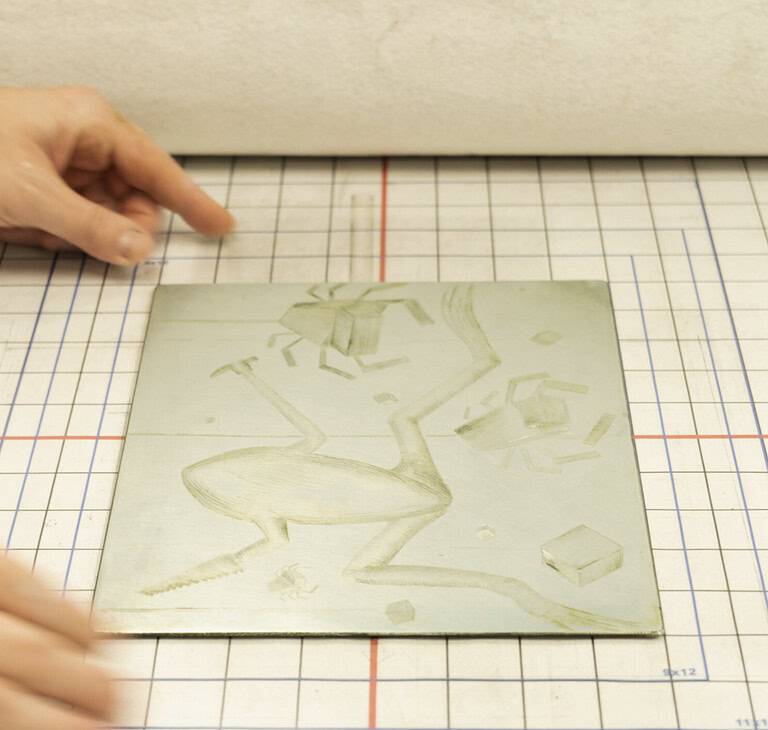 A person holds a linocut plate with an engraved abstract frog design combining geometric shapes and organic shapes, on top a grid-lined workspace.