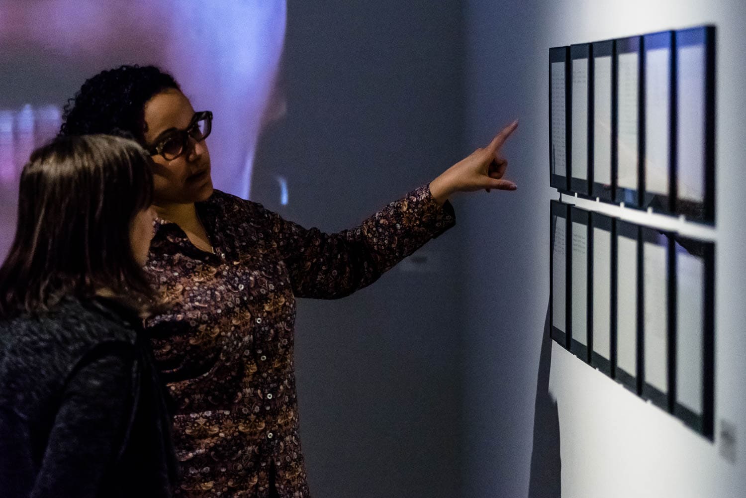 Two people stand in a dimly lit gallery; one points at a series of framed artworks on the wall while explaining something to the other.