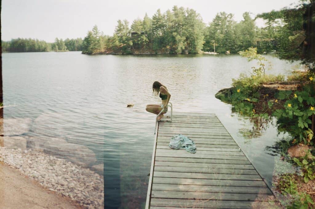 A photograph of person in a swimsuit sitting at the edge of a wooden dock extending into a calm lake, surrounded by trees.