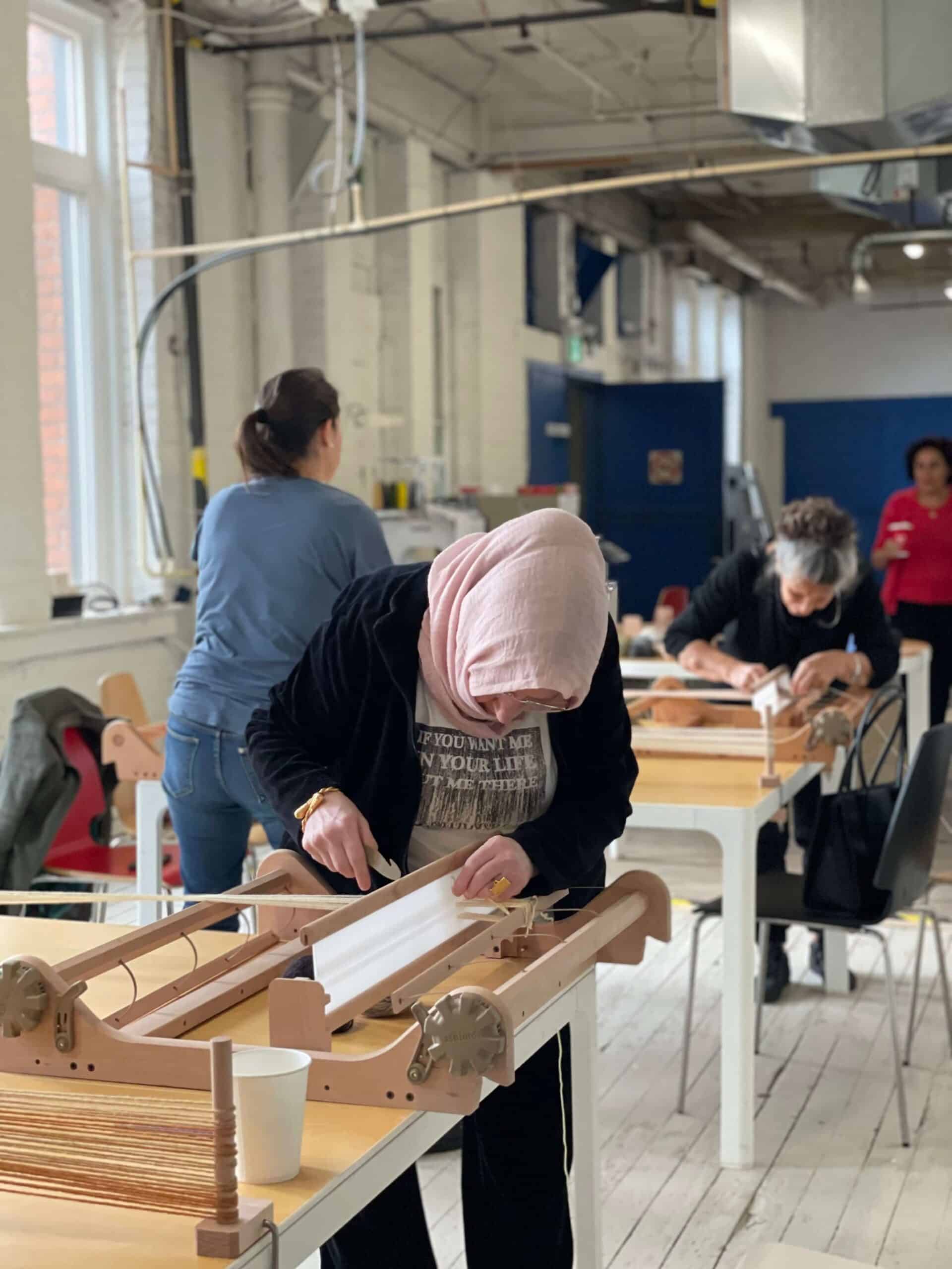 Several people work at weaving looms in a bright, spacious studio with large windows and white floors. One person in the foreground wears a pink headscarf and focuses on their loom.