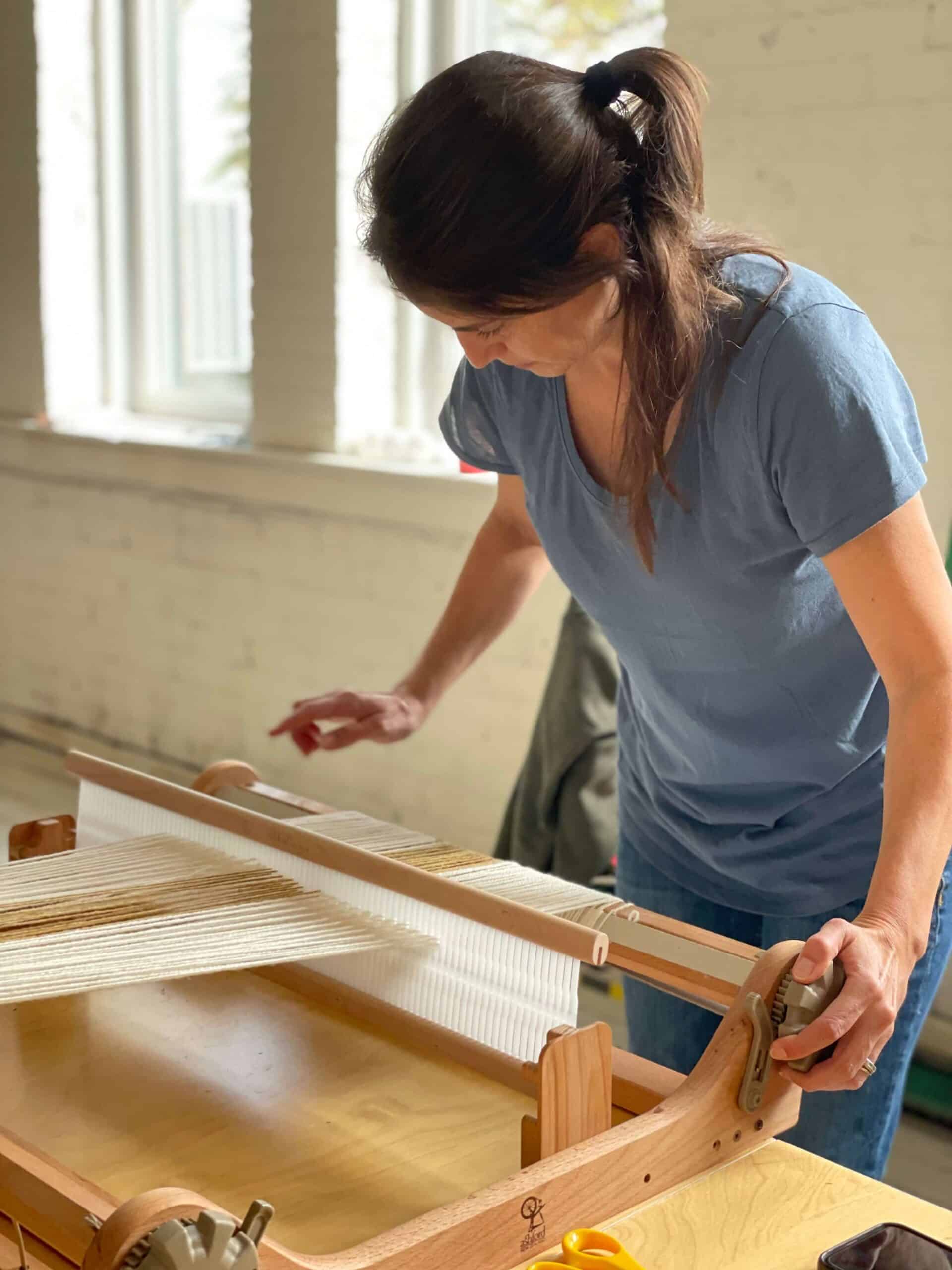A participant weaving white and tan threads on a wooden loom in a bright studio workshop.