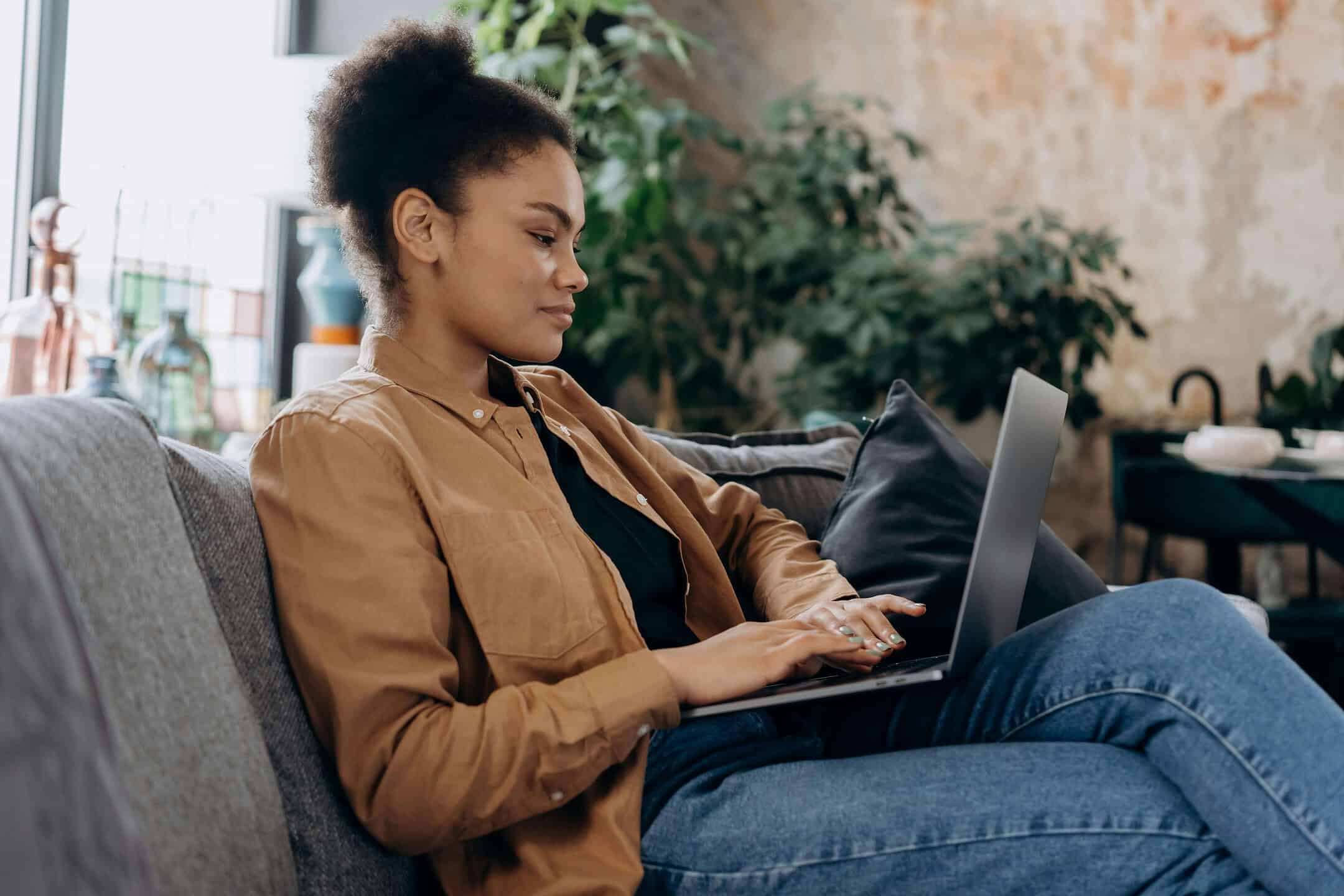 A woman in a tan button-up shirt sits on a grey couch, drafting a document on her laptop. She appears focused and relaxed, with plants and home decor visible in the background.