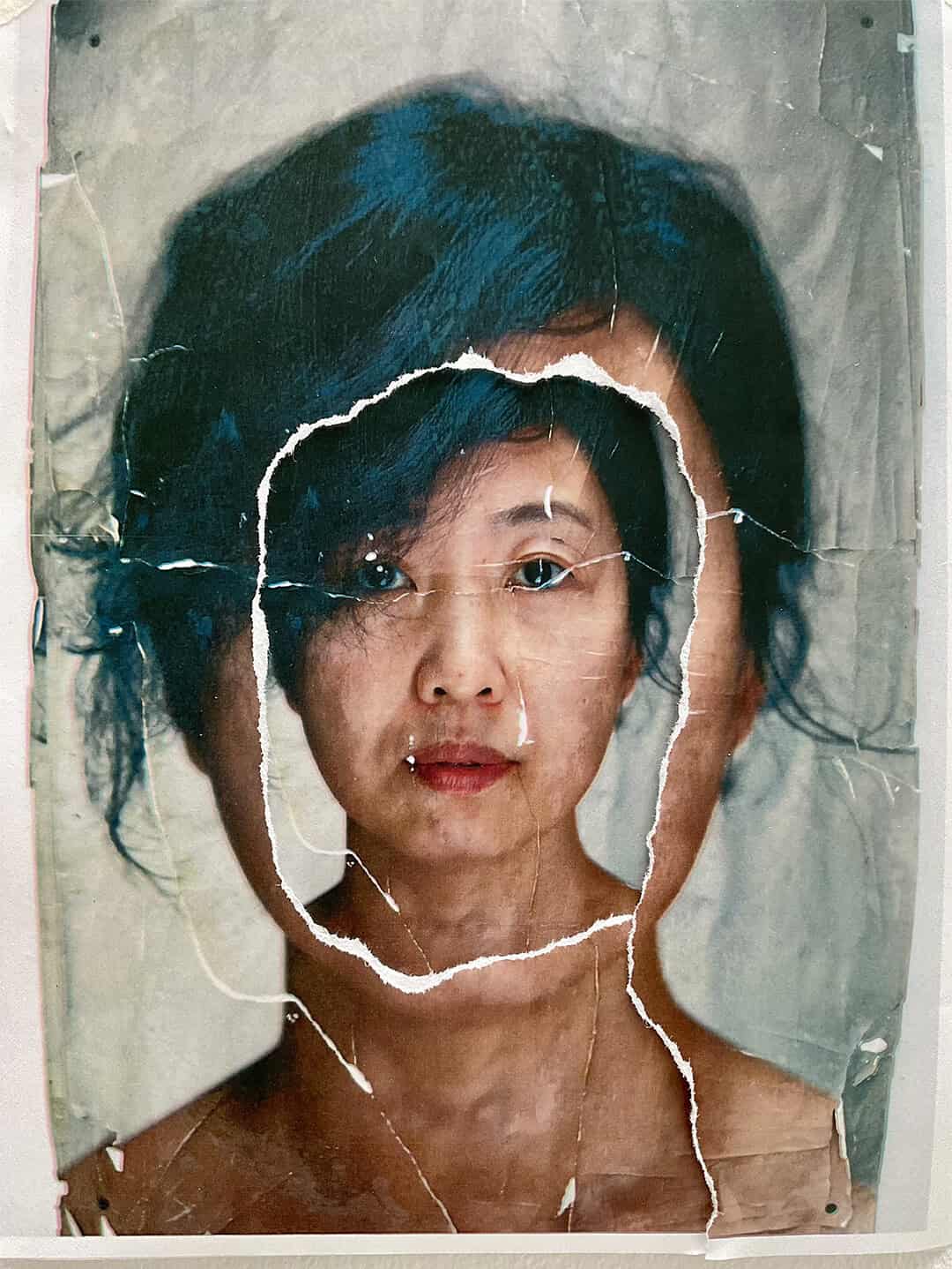 A portrait of a woman with dark hair is layered over a similar portrait, with the top image torn to reveal her face beneath, evoking shifting borders and creating a surreal, double-exposure effect. The edges of the tear are rough and uneven.