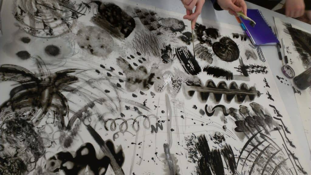Two people point at a large tabletop canvas covered in black ink doodles, smudges, swirls, and abstract patterns. A purple notebook and marker are visible in the corner.