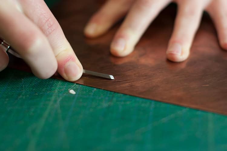 A close-up of a person using a metal tool to carve or etch a sheet of copper, with fingers holding the material steady on a green cutting mat.