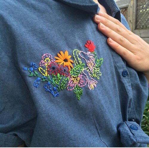 A person wearing a blue denim shirt with colourful embroidered flowers places their hand over their chest.