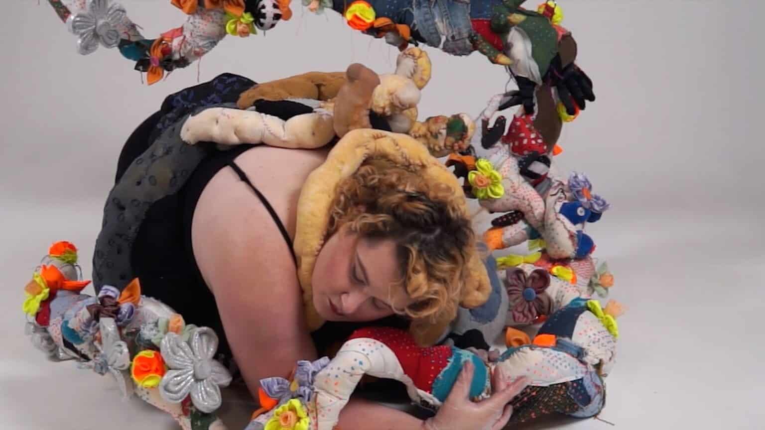A person with curly blonde hair wearing a black outfit is curled up on the floor, surrounded by colourful, stuffed fabric shapes and soft sculptures, all set against a plain white background.