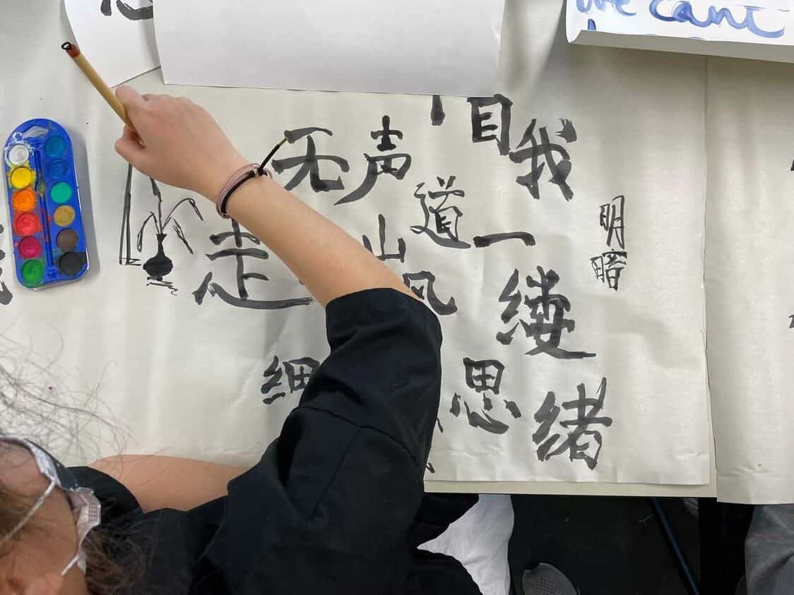 A person practices calligraphy, painting black Chinese characters on white paper. Nearby, a watercolour paint set and brush rest on the table. The person wears a dark shirt.