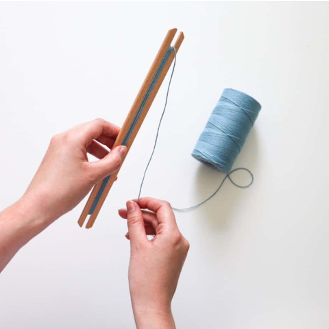 A person begins weaving by wrapping light blue string around two parallel wooden sticks, with a spool of the same string nearby on a white surface.